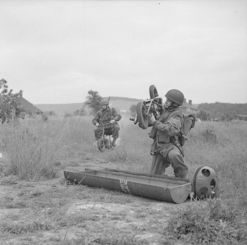 A paratrooper unloads a Welbike from its container, while another paratrooper rides past